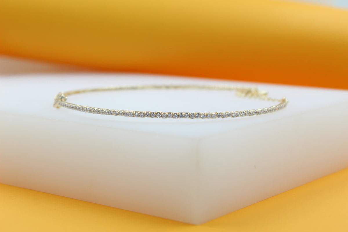A delicate gold tennis bracelet encrusted with sparkling cubic zirconia stones rests elegantly on a white display block against a warm yellow background.