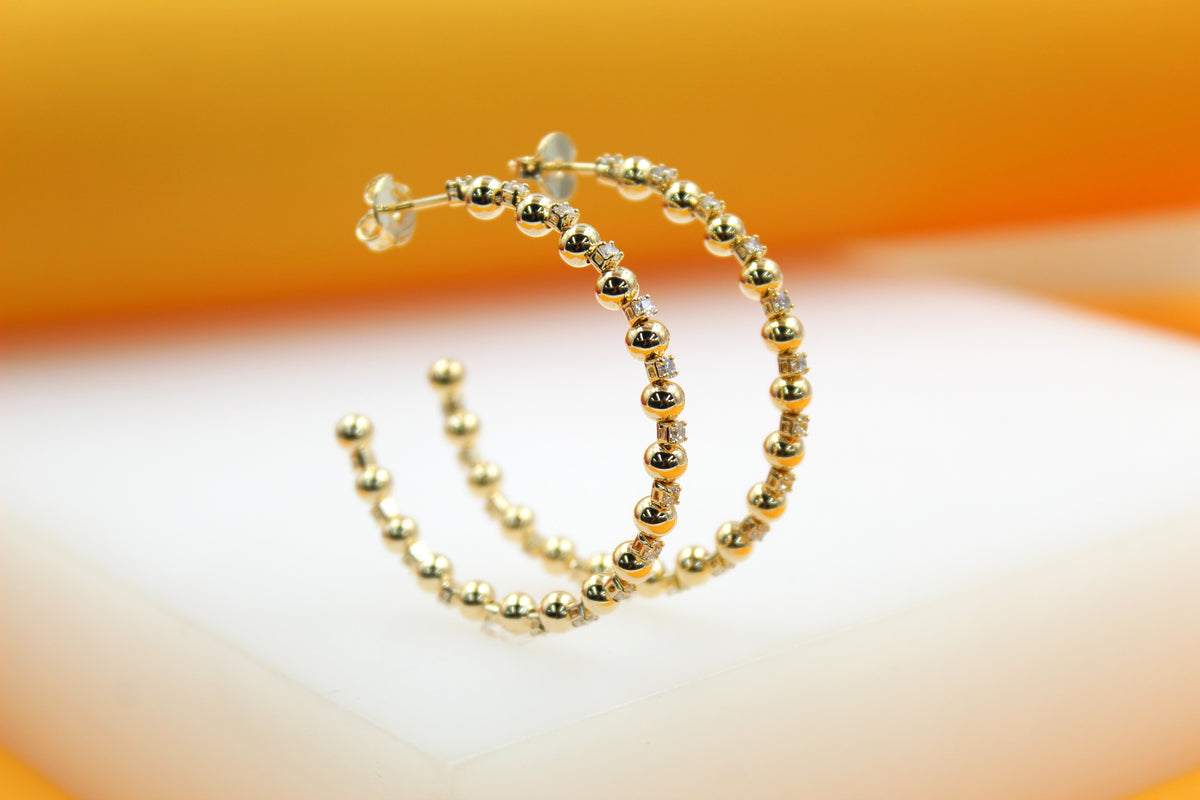 Two elegant gold hoop earrings featuring a beaded texture and sparkling stone accents rest on a white display surface against a warm orange backdrop.