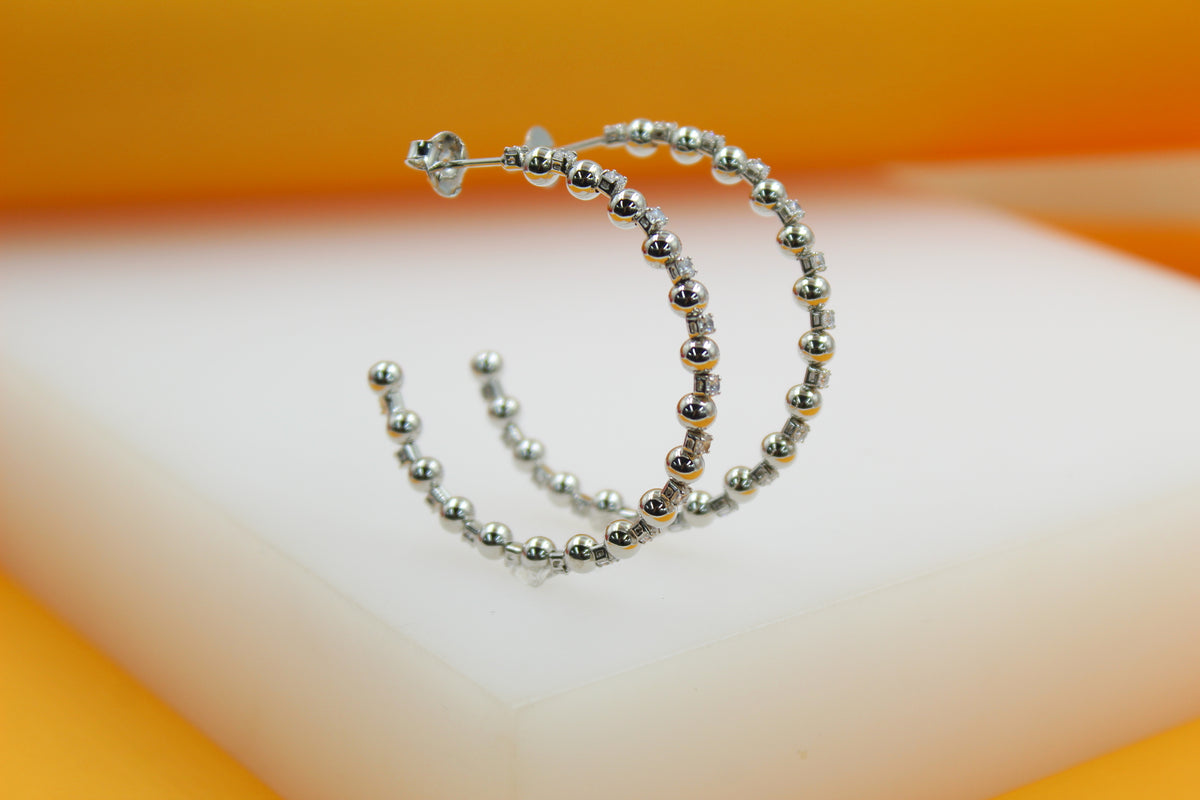 A pair of sterling silver hoop earrings adorned with alternating beads and sparkling cubic zirconia stones rests on a white display surface against a warm orange backdrop.