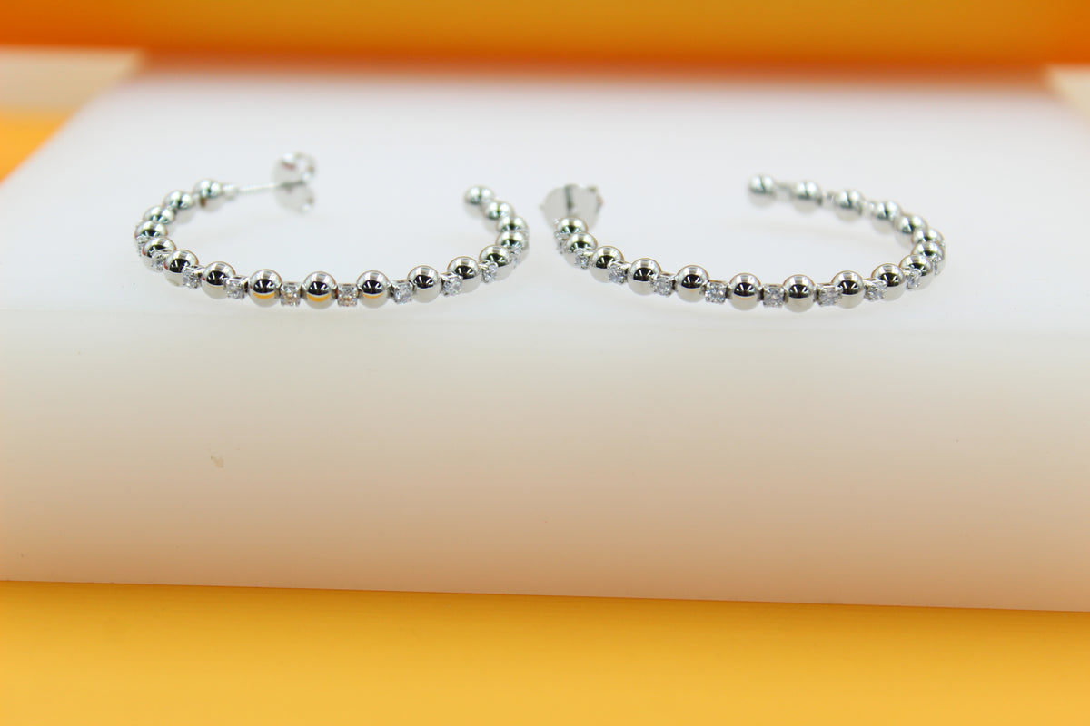 A pair of silver hoop earrings adorned with alternating beads and sparkling gemstones rests on a white display surface against a vibrant orange backdrop.