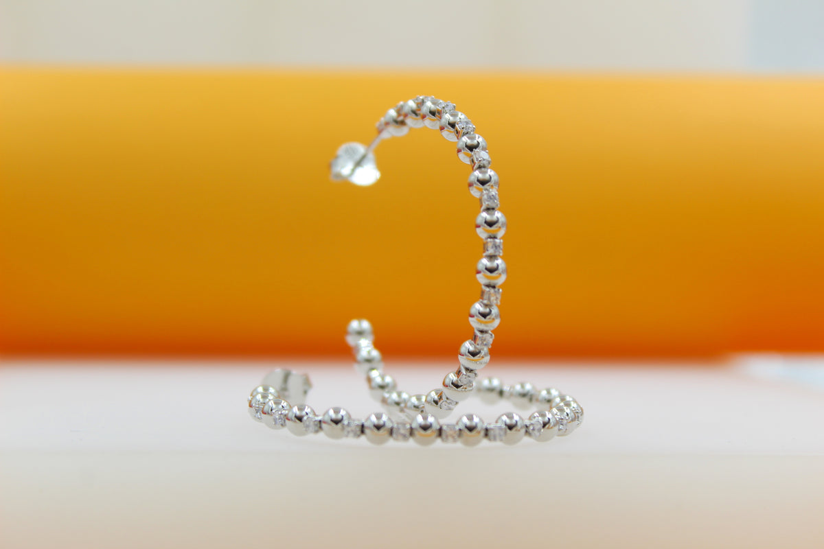 A pair of sparkling silver hoop earrings rest on a reflective surface against a vibrant orange and white background.