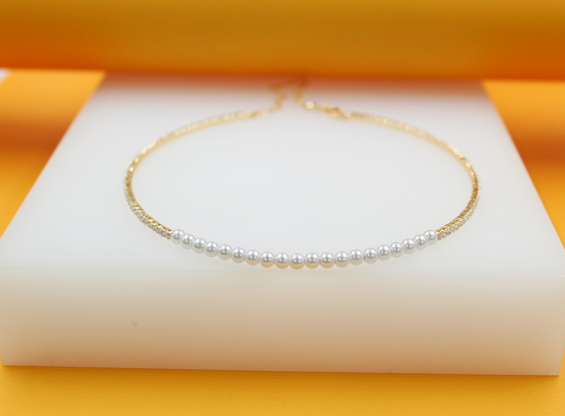 A delicate gold pearl choker necklace featuring a row of white pearls rests on a white display block against an orange backdrop.