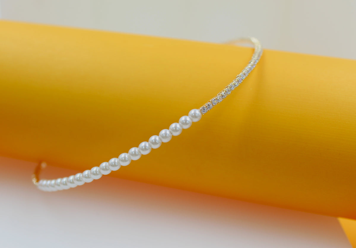 A delicate necklace featuring a row of white pearls transitioning into a sparkly diamond-encrusted band rests against a bright yellow cylinder.