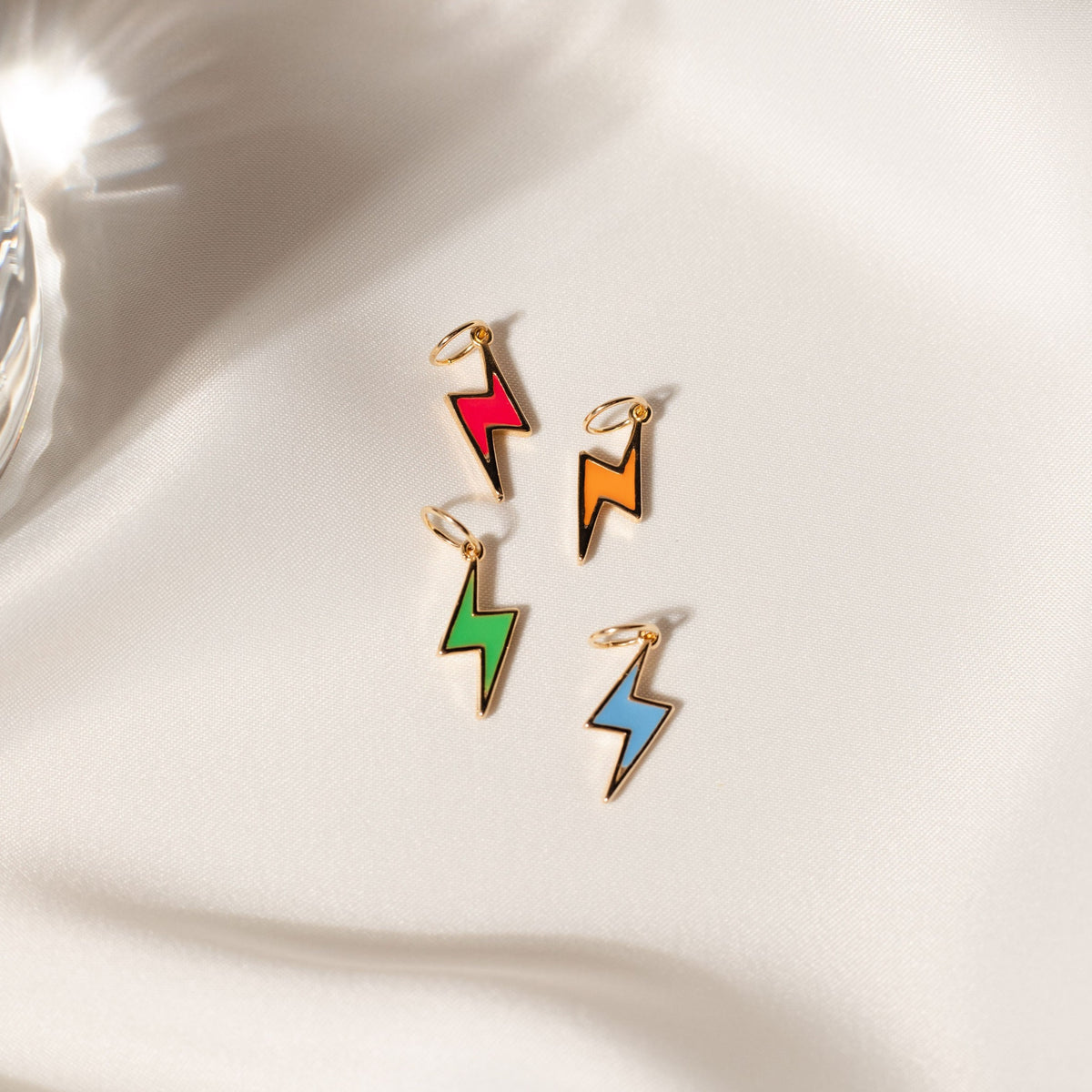 Four colorful enamel lightning bolt charms in red, orange, green, and blue rest on a white silk background.