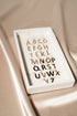 A white tray holds an alphabet set of gold initial letter charms, each featuring a small jump ring, displayed on a satin fabric background.