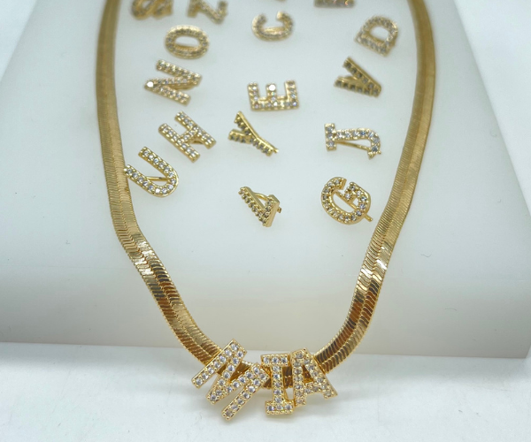 A gold herringbone chain necklace spells out the name MIA with diamond-encrusted letter charms, surrounded by scattered alphabet pendants.