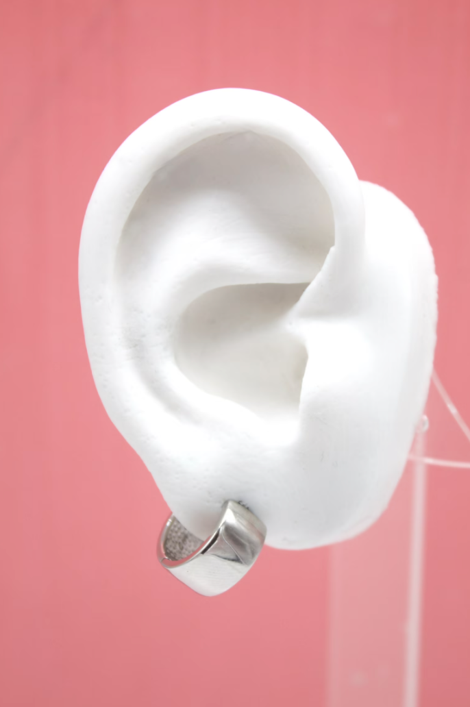 A white mannequin ear displays a chunky silver huggie hoop earring against a soft pink background.