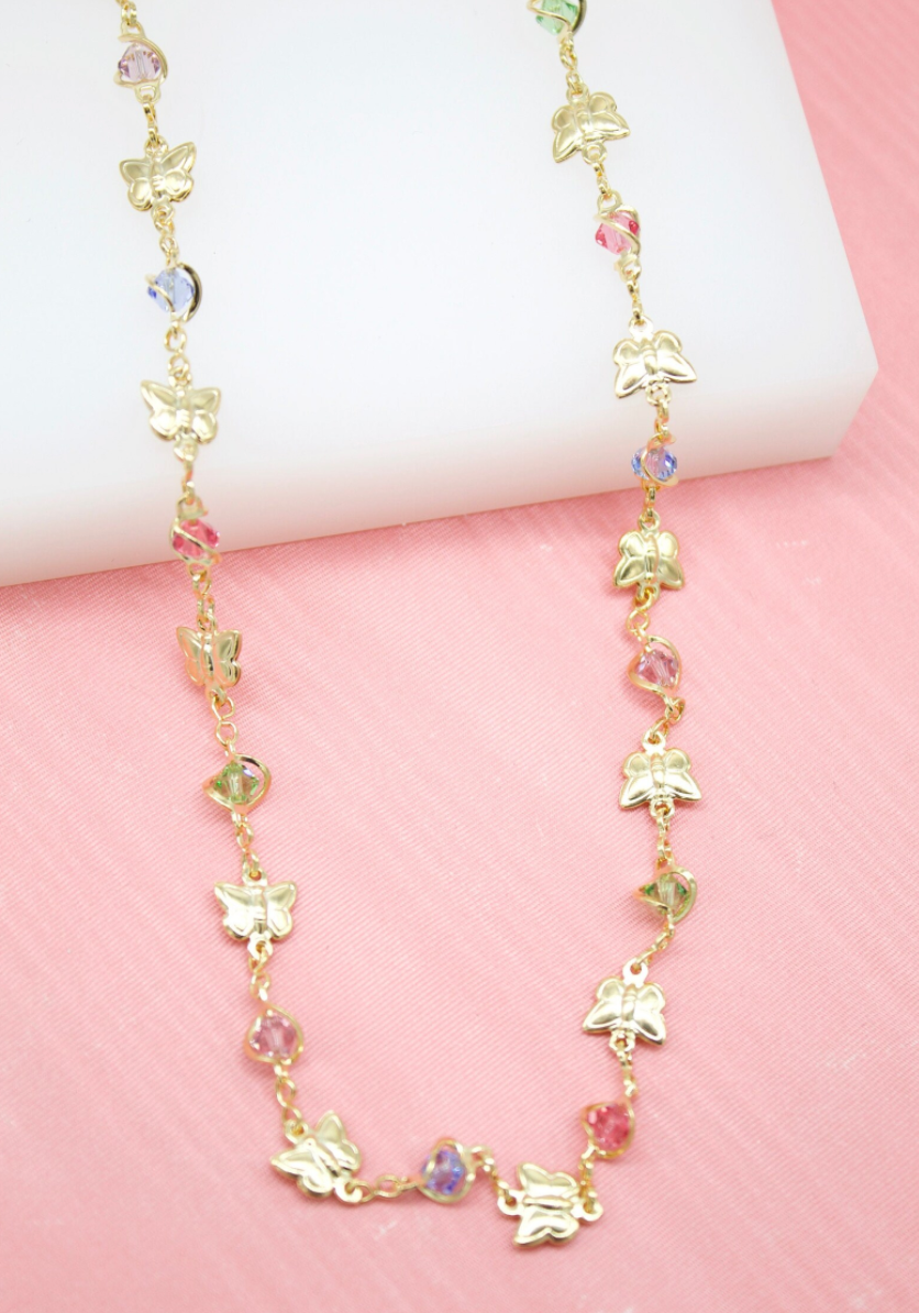 A delicate gold butterfly necklace with colorful crystal beads rests against a pink and white display background.