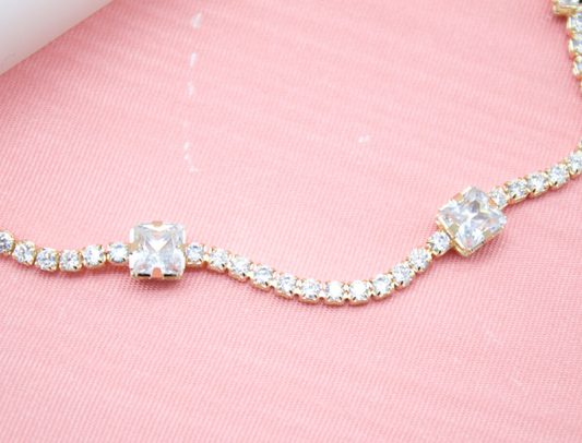 A gold tennis necklace sparkles against a soft pink background, featuring rows of small round crystals accented by larger square cubic zirconia stones.