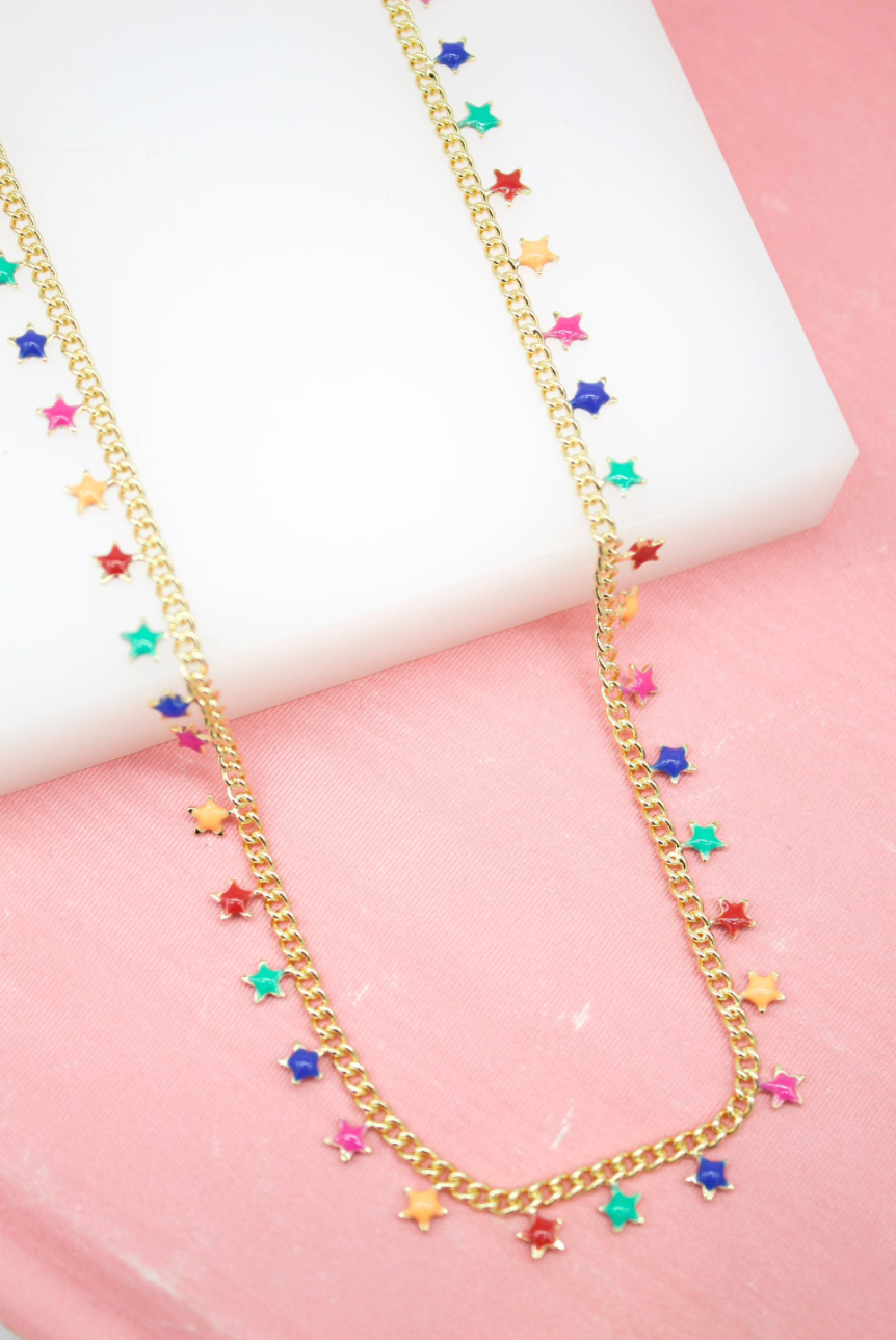 A delicate gold chain necklace with colorful enamel star charms rests on a white block against a pink background.