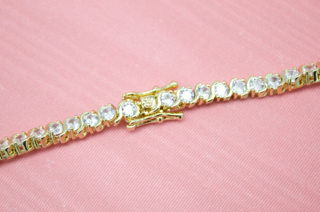 A gold tennis bracelet featuring sparkling cubic zirconia stones rests diagonally against a soft pink textured background, highlighting its intricate box clasp.
