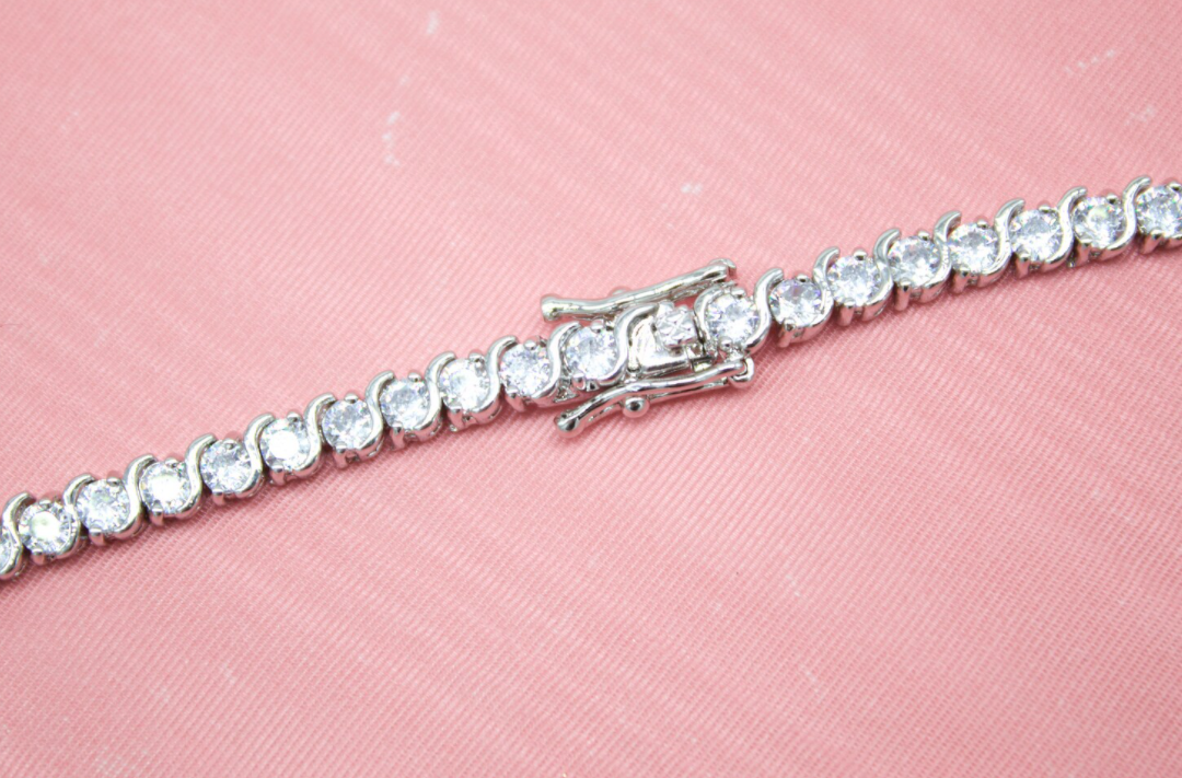 A sparkling silver tennis bracelet with a secure box clasp lies diagonally across a textured pink background.