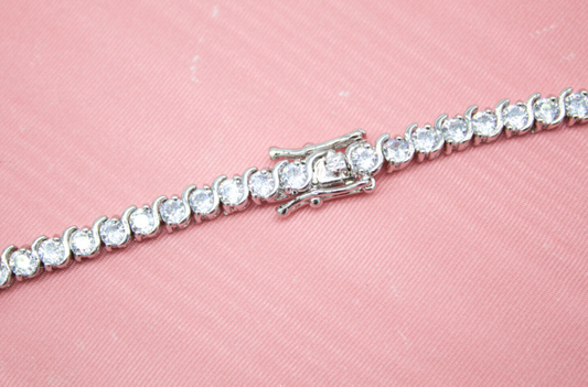 A sparkling silver tennis bracelet with a secure box clasp lies diagonally across a textured pink background.