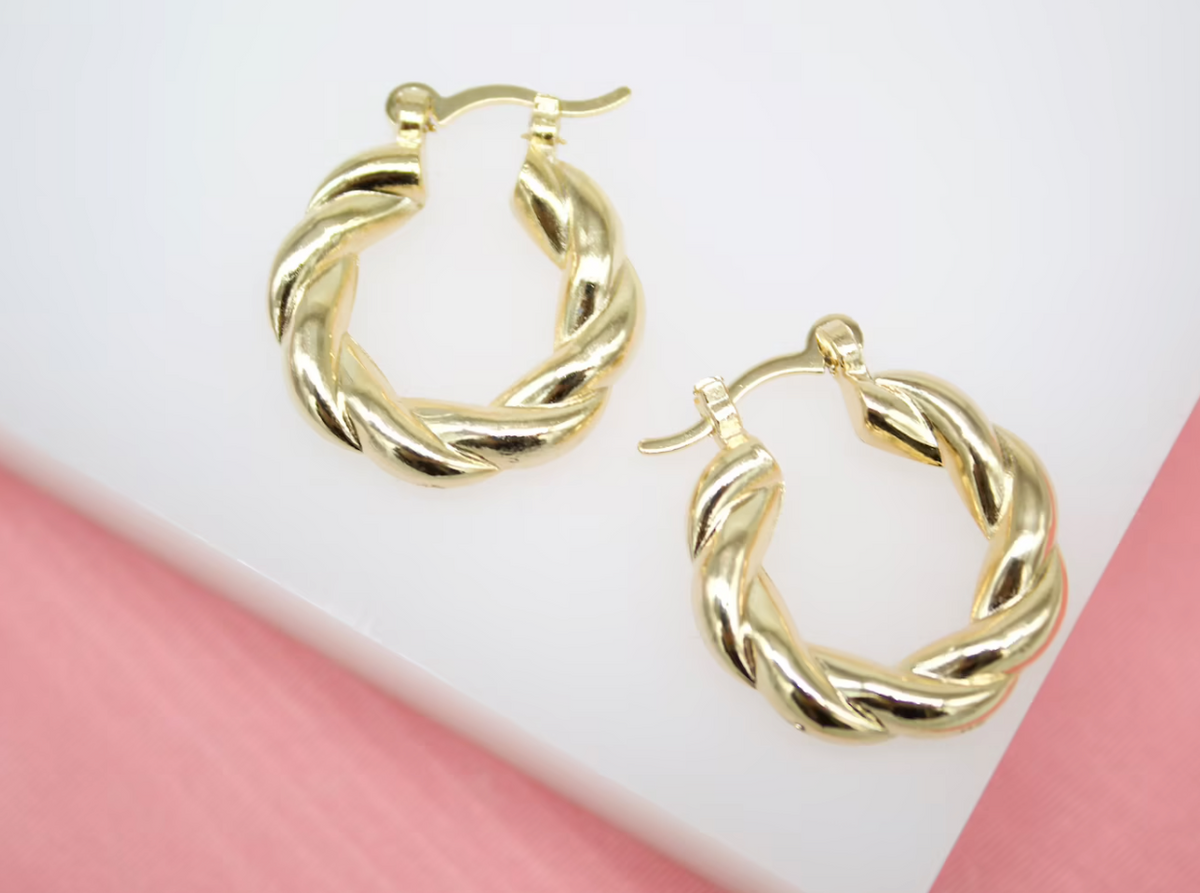 A pair of polished gold twist hoop earrings rests against a white and pink display surface.