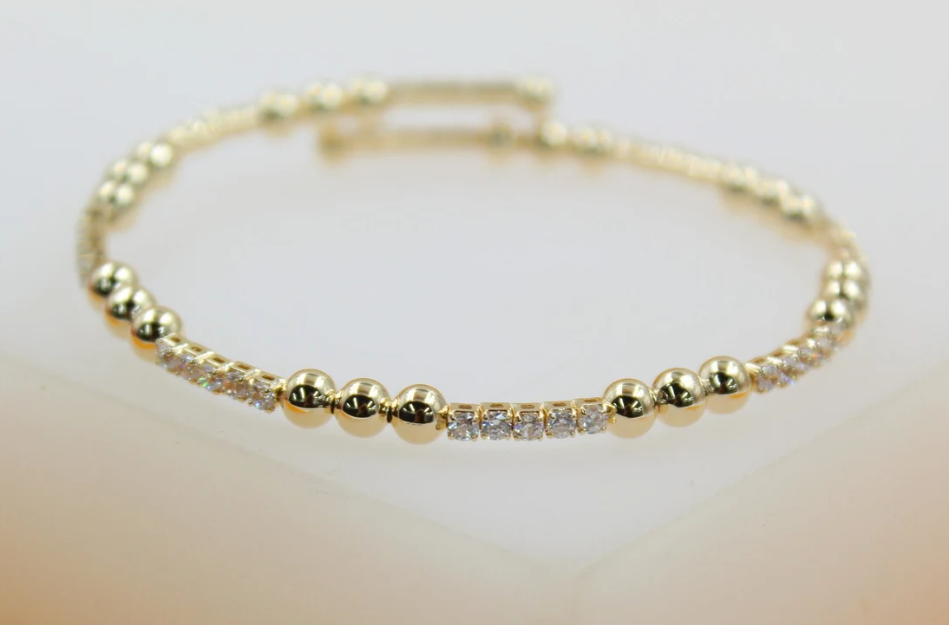 A delicate gold beaded bracelet with alternating sections of polished gold spheres and sparkling cubic zirconia stones rests on a light surface.