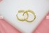 Two interlocking gold hoop earrings featuring a textured, lattice design rest on a white surface against a pink background.