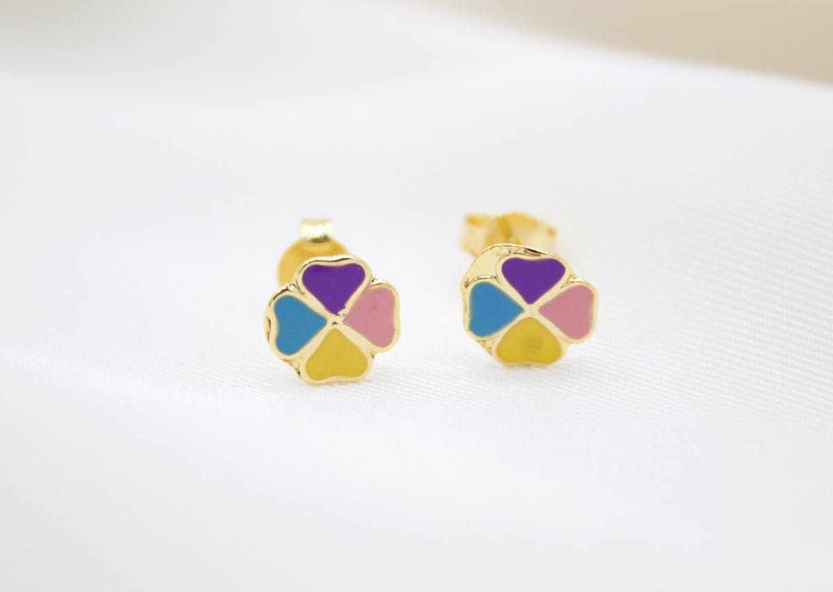 A pair of gold stud earrings shaped like four-leaf clovers features colorful enamel petals in purple, blue, yellow, and pink on a white fabric background.