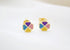A pair of gold stud earrings shaped like four-leaf clovers features colorful enamel petals in purple, blue, yellow, and pink on a white fabric background.