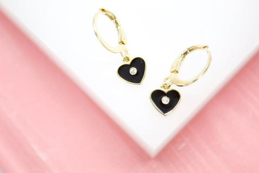 A pair of gold huggie hoop earrings featuring black enamel heart charms with center diamonds rests on a white display against a soft pink background.
