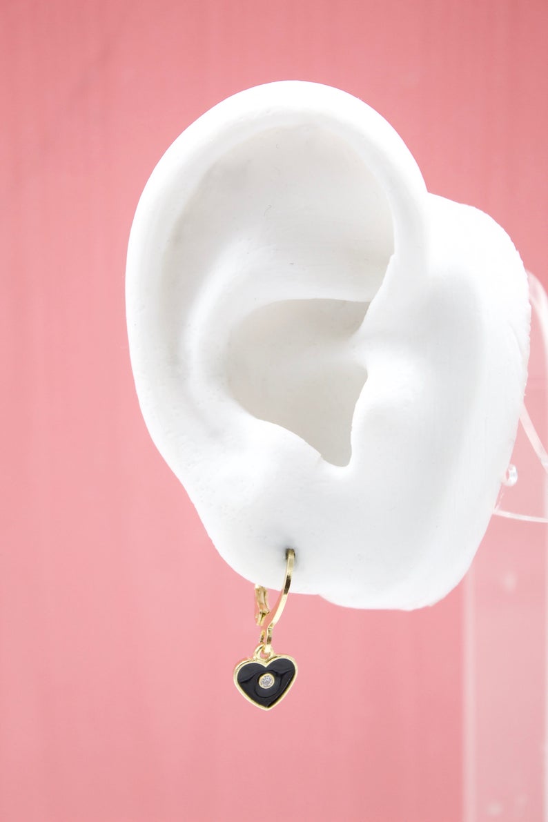 A white mannequin ear displays a gold huggie hoop earring featuring a dangling black enamel heart charm with a tiny center diamond.