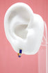 A white model ear displays a small gold hoop earring featuring vibrant blue enamel and a sparkling diamond accent.