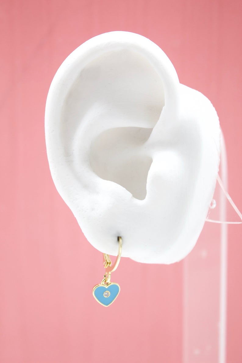 A white mannequin ear displays a gold huggie hoop earring featuring a bright blue enamel heart charm with a small central diamond.