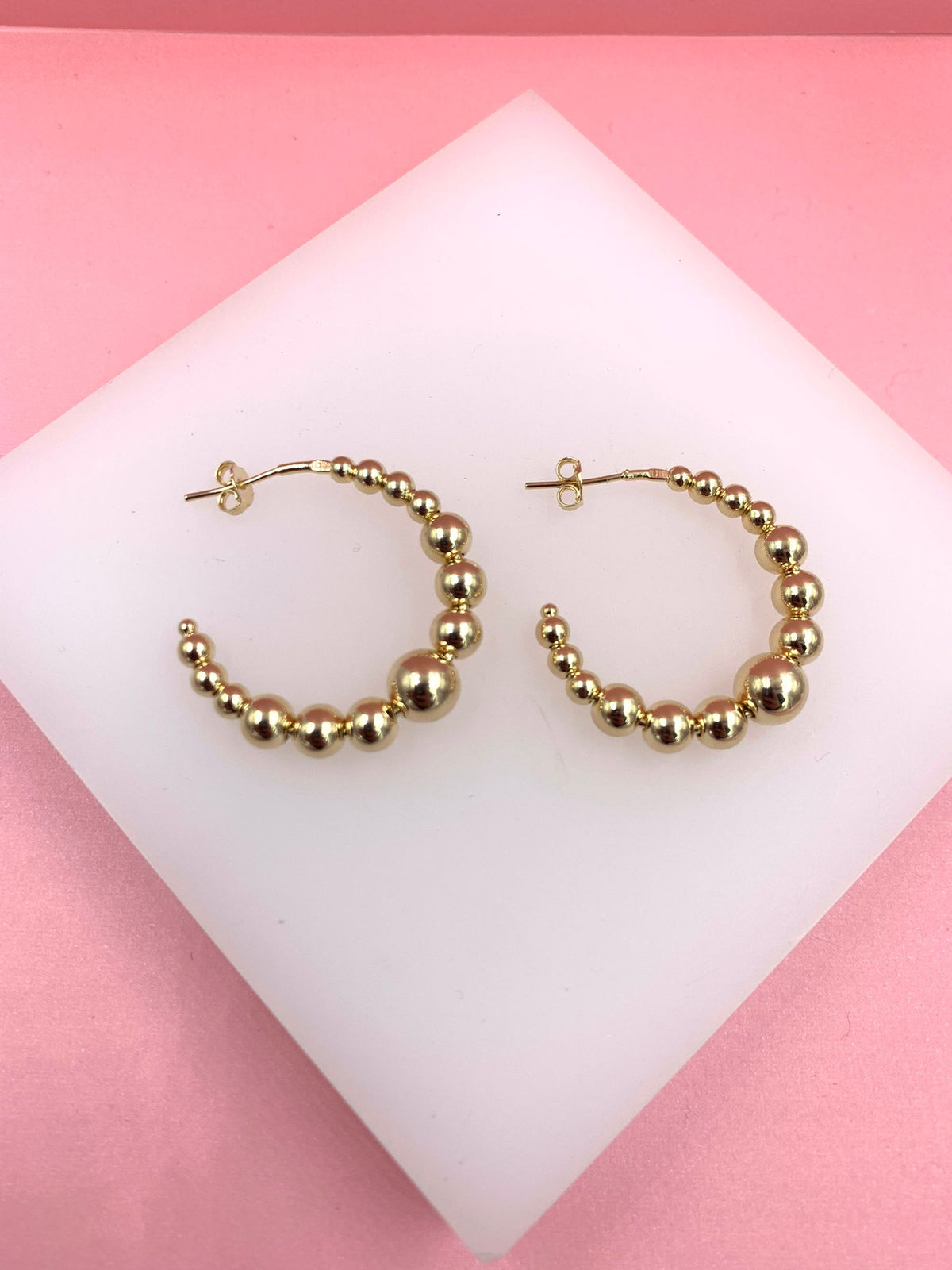 A pair of gold ball hoop earrings rests on a white display against a pink background.
