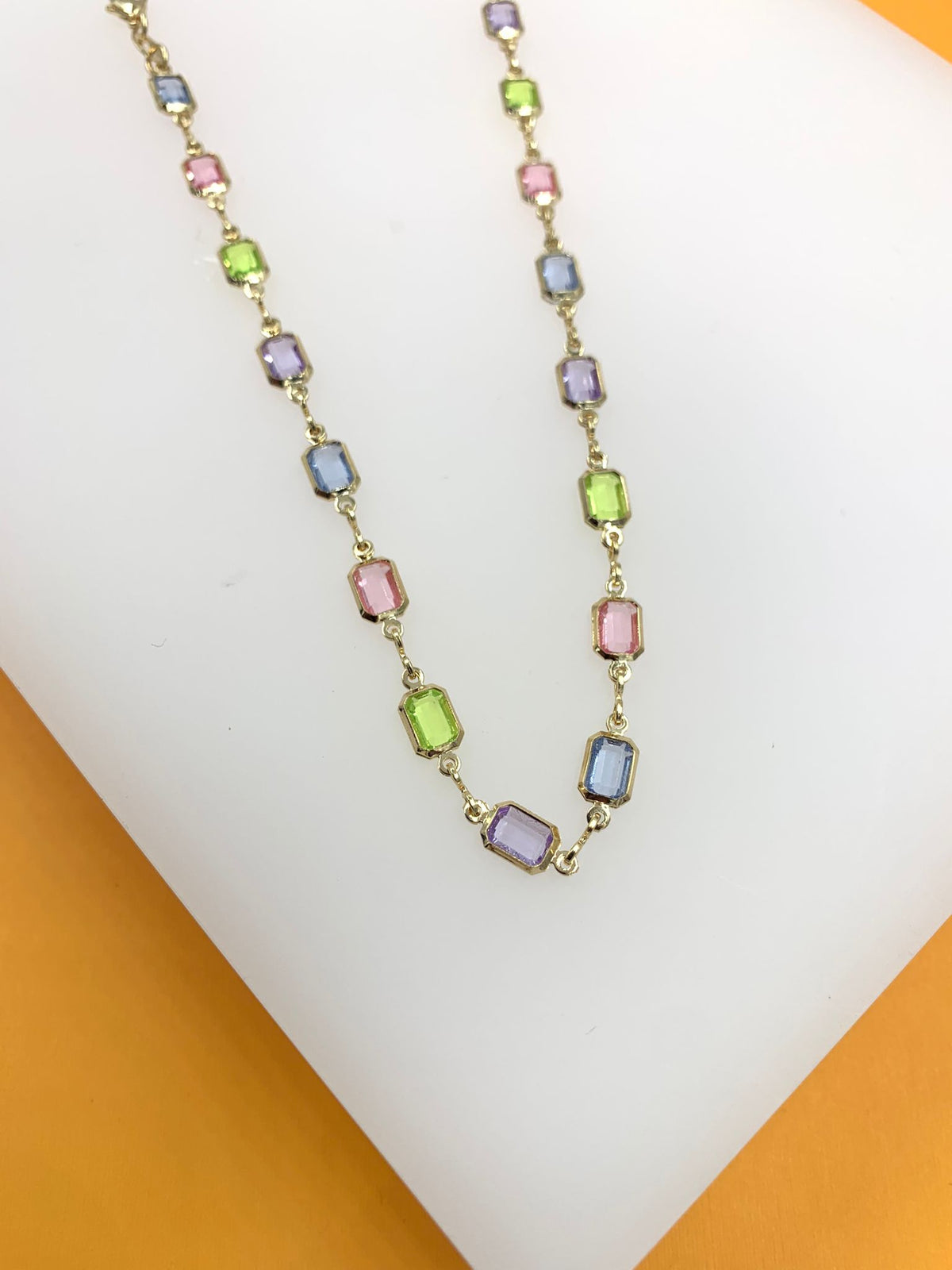 A delicate gold chain necklace drapes diagonally across a white display, featuring vibrant multi-colored gemstone stations in shades of pink, green, blue, and purple.