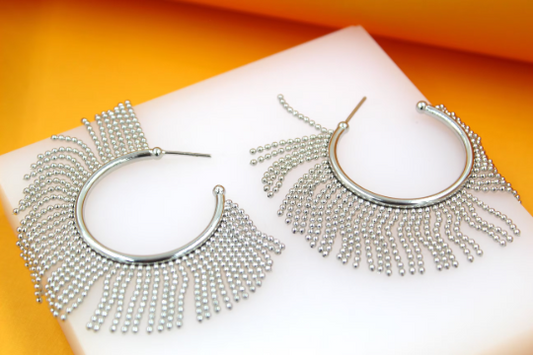 A pair of silver hoop earrings adorned with dangling beaded fringe rests on a white display surface against a vibrant orange background.