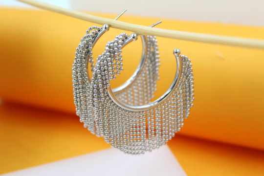 A pair of silver hoop earrings adorned with beaded fringe hangs delicately from a wooden stick against a bright yellow background.