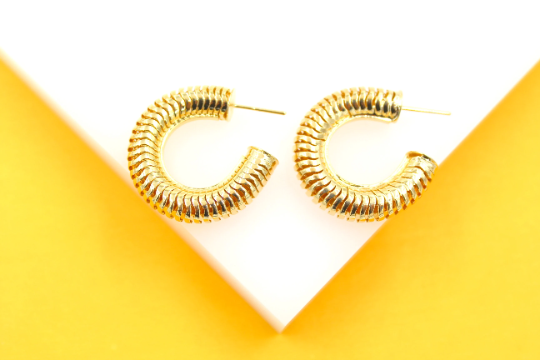 A pair of gold-plated, ribbed hoop earrings rests on a white and yellow background.
