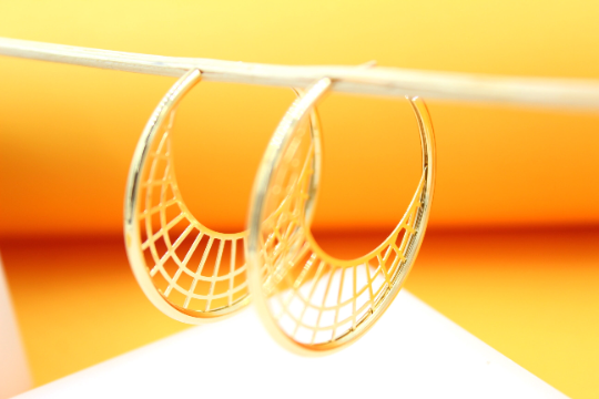 A pair of gold hoop earrings with an intricate mesh pattern hang delicately against a bright yellow background.