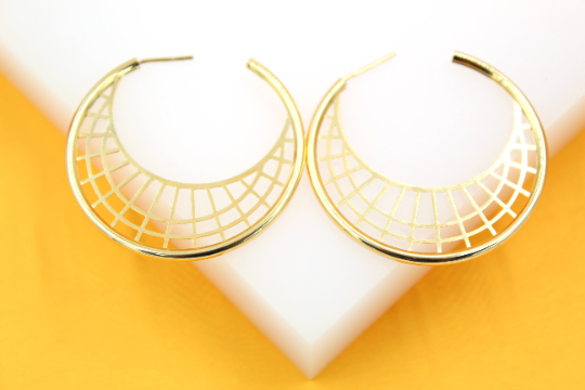 A pair of large, gold-plated hoop earrings with intricate geometric mesh patterns rests on a white display against a vibrant yellow background.