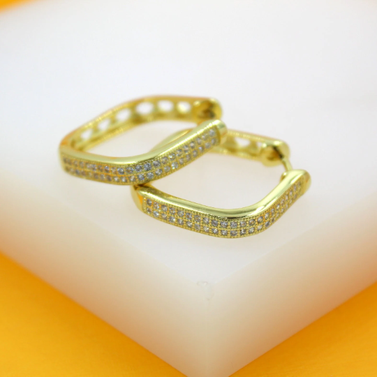 A pair of geometric gold huggie hoop earrings encrusted with pavé diamonds rests on a white display block.