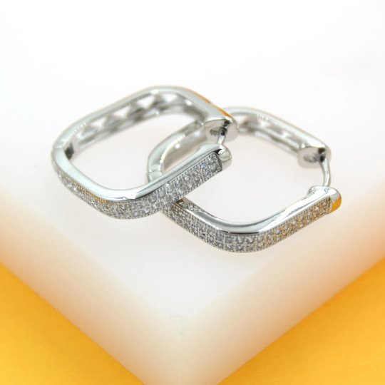 A pair of silver square hoop earrings encrusted with sparkling cubic zirconia rest on a white surface against an orange background.