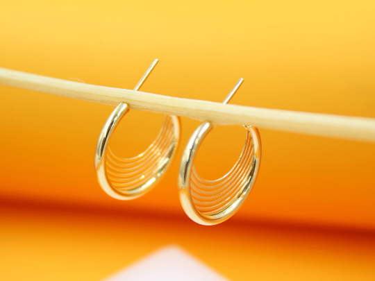 A pair of polished gold multi-strand hoop earrings hangs gracefully from a wooden stick against a warm orange background.