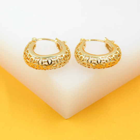 A pair of intricate gold filigree hoop earrings rests on a white and yellow display block.
