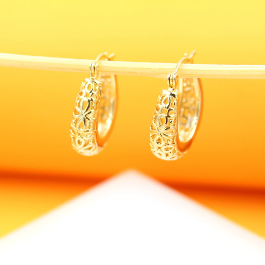 A pair of intricate gold filigree hoop earrings hangs delicately from a wooden stick against a bright yellow background.