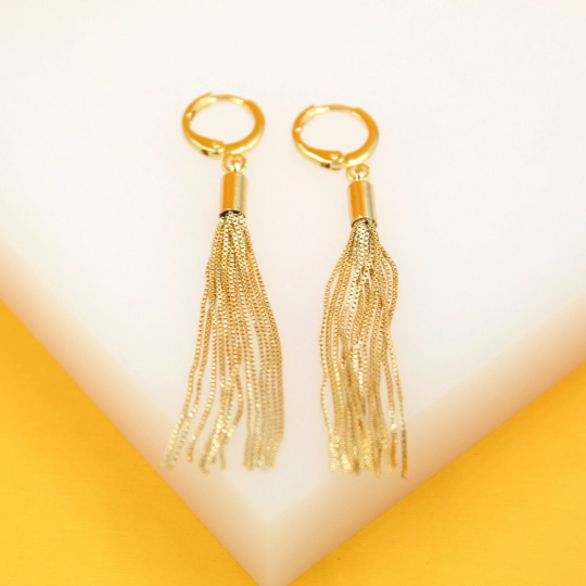 A pair of elegant gold tassel earrings resting on a white display surface against a vibrant yellow background.
