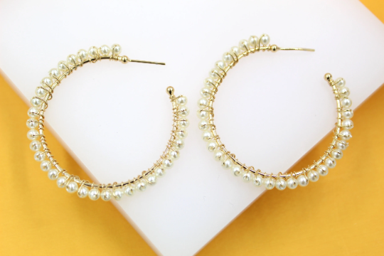 A pair of elegant gold hoop earrings embellished with delicate white faux pearls resting on a white display against a vibrant yellow background.