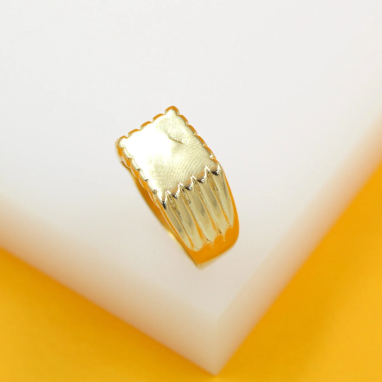 A gold signet ring with a textured, rectangular face rests on a white block against a yellow background.