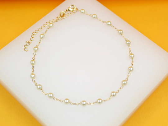A delicate gold anklet features evenly spaced white faux pearls resting gracefully on a white display block against a vibrant yellow background.