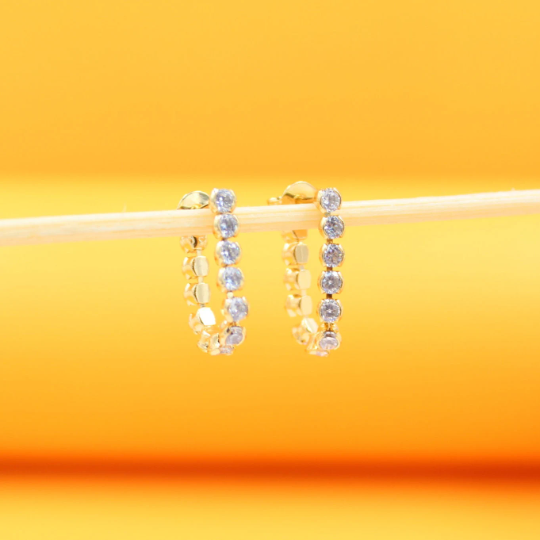A pair of gold hoop earrings embellished with sparkling cubic zirconia stones rests horizontally on a wooden stick against a bright yellow background.