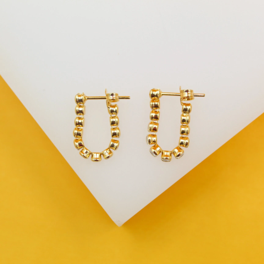 Two U-shaped gold hoop earrings featuring sparkling pave-set stones rest on a white display surface against a yellow background.