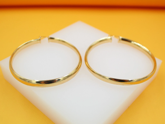 A pair of polished gold hoop earrings rests on a white display block against a vibrant yellow background.
