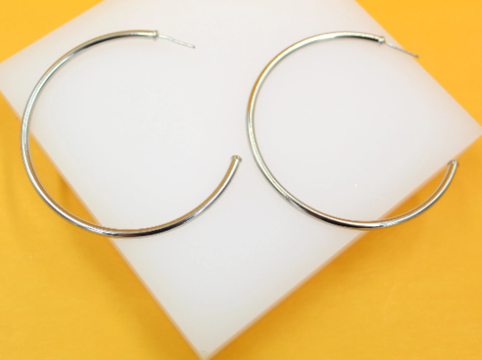 A pair of large silver hoop earrings rests on a white display block against a vibrant yellow background.