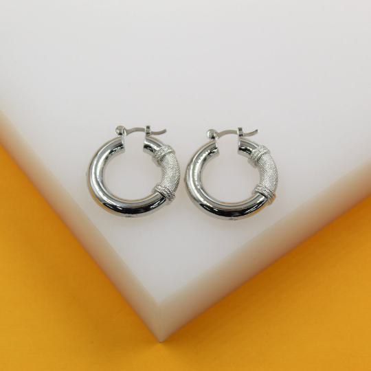 A pair of thick silver hoop earrings featuring textured mesh details rest neatly on a white and yellow display stand.