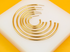 A graduated set of polished gold-plated hoop earrings rests on a white display stand against a vibrant yellow background.