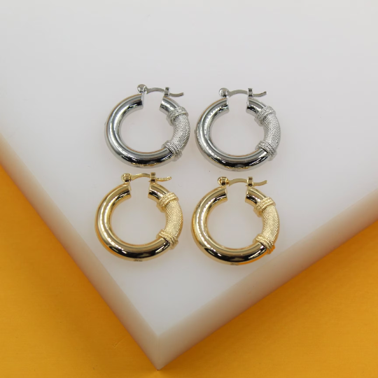 Two pairs of chunky hoop earrings—one silver and one gold, featuring a textured, woven section—rest on a white display block.