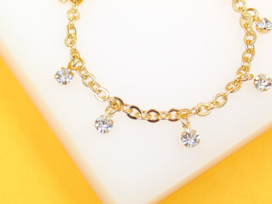 A delicate gold chain bracelet features sparkling rhinestone charms hanging against a white and yellow background.